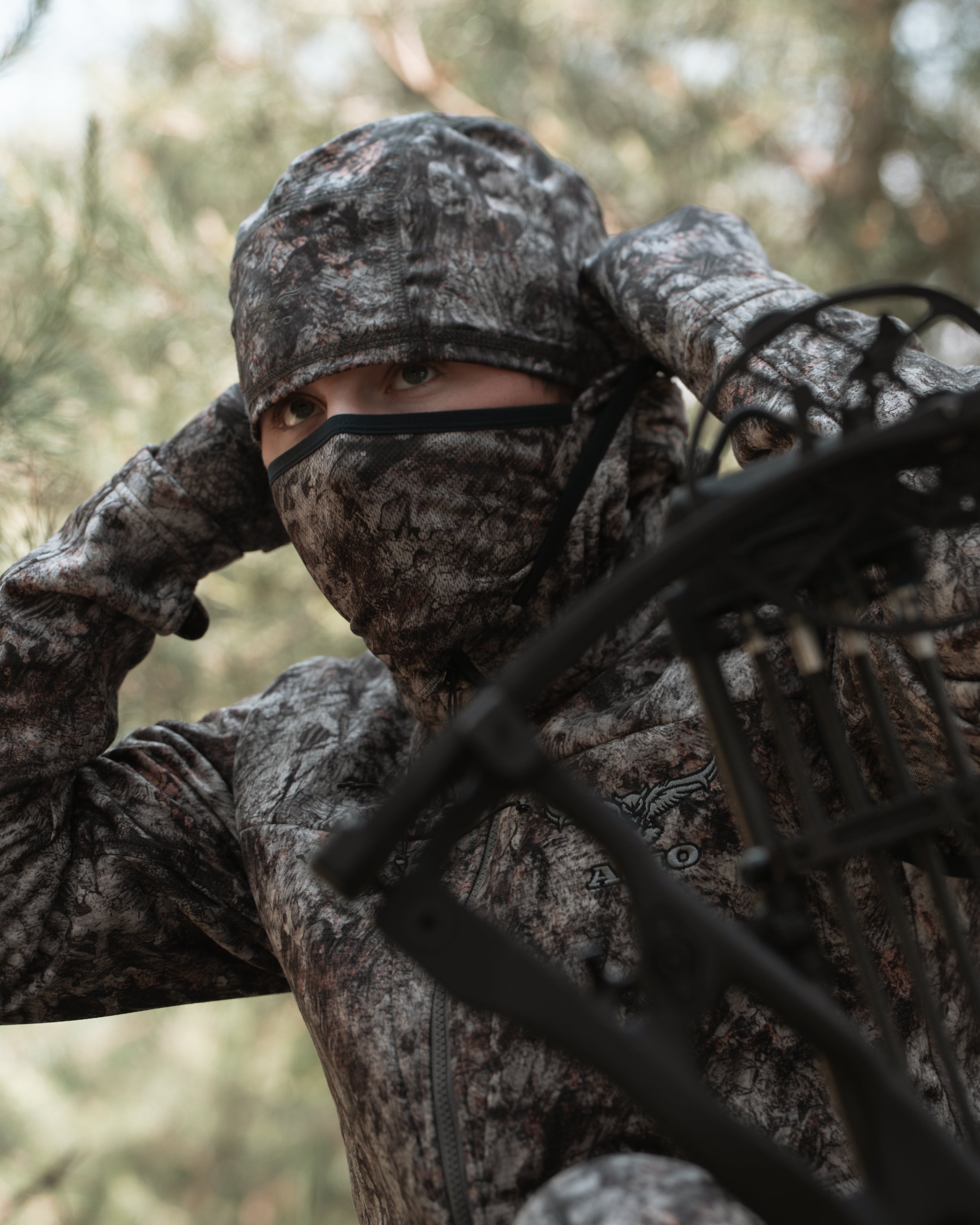 Bowhunter in RAPTOR camo midweight hoodie adjusting built-in face mask in pine woods.