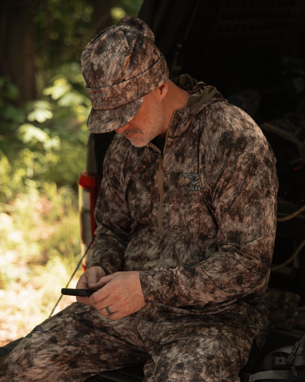 Hunter wearing ASIO Gear RAPTOR camo fleece-lined billed beanie and mid-season system while checking phone at tailgate