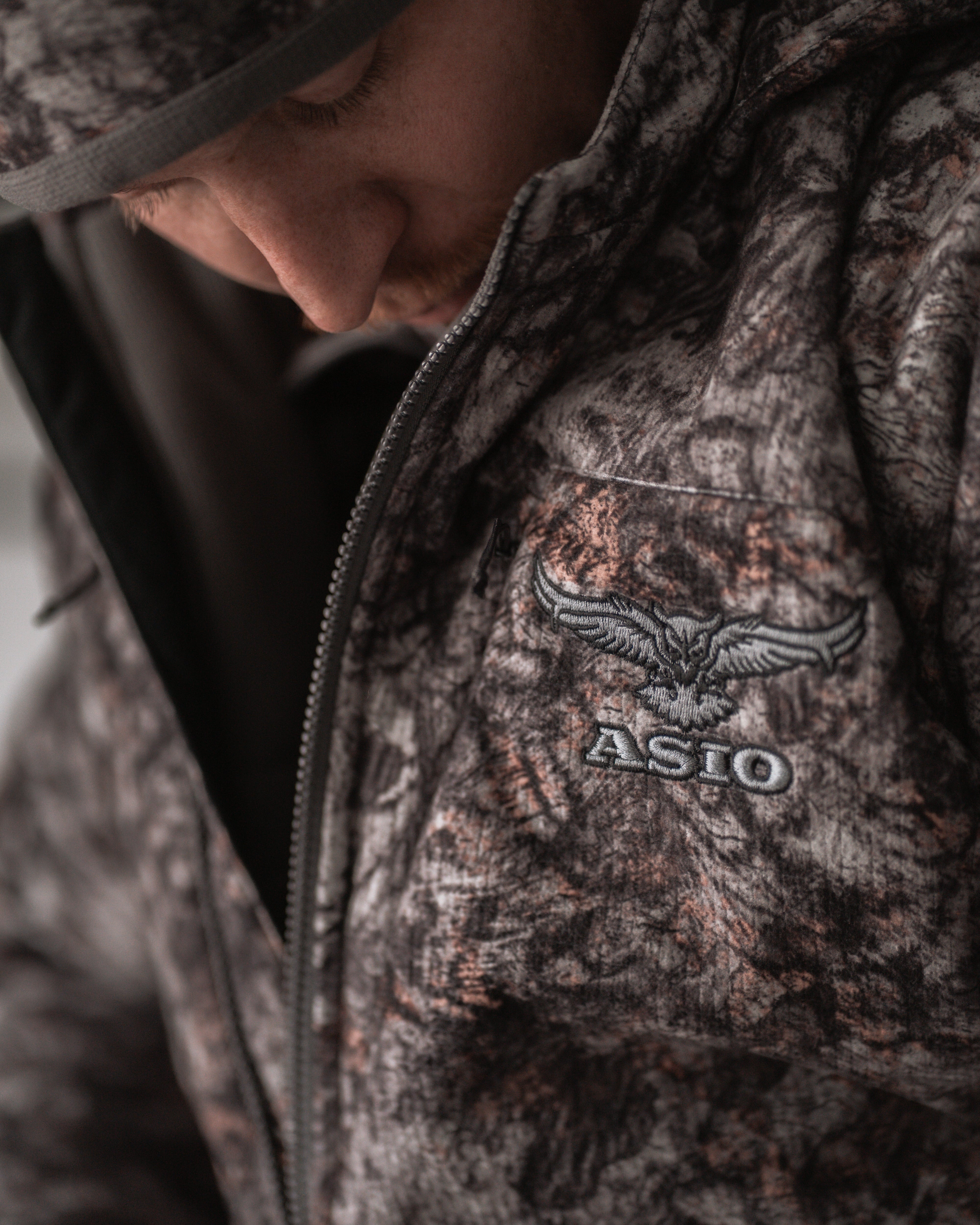 Close-up of ASIO Gear RAPTOR camo heavyweight hooded jacket showing logo embroidery, zipper, and fabric texture