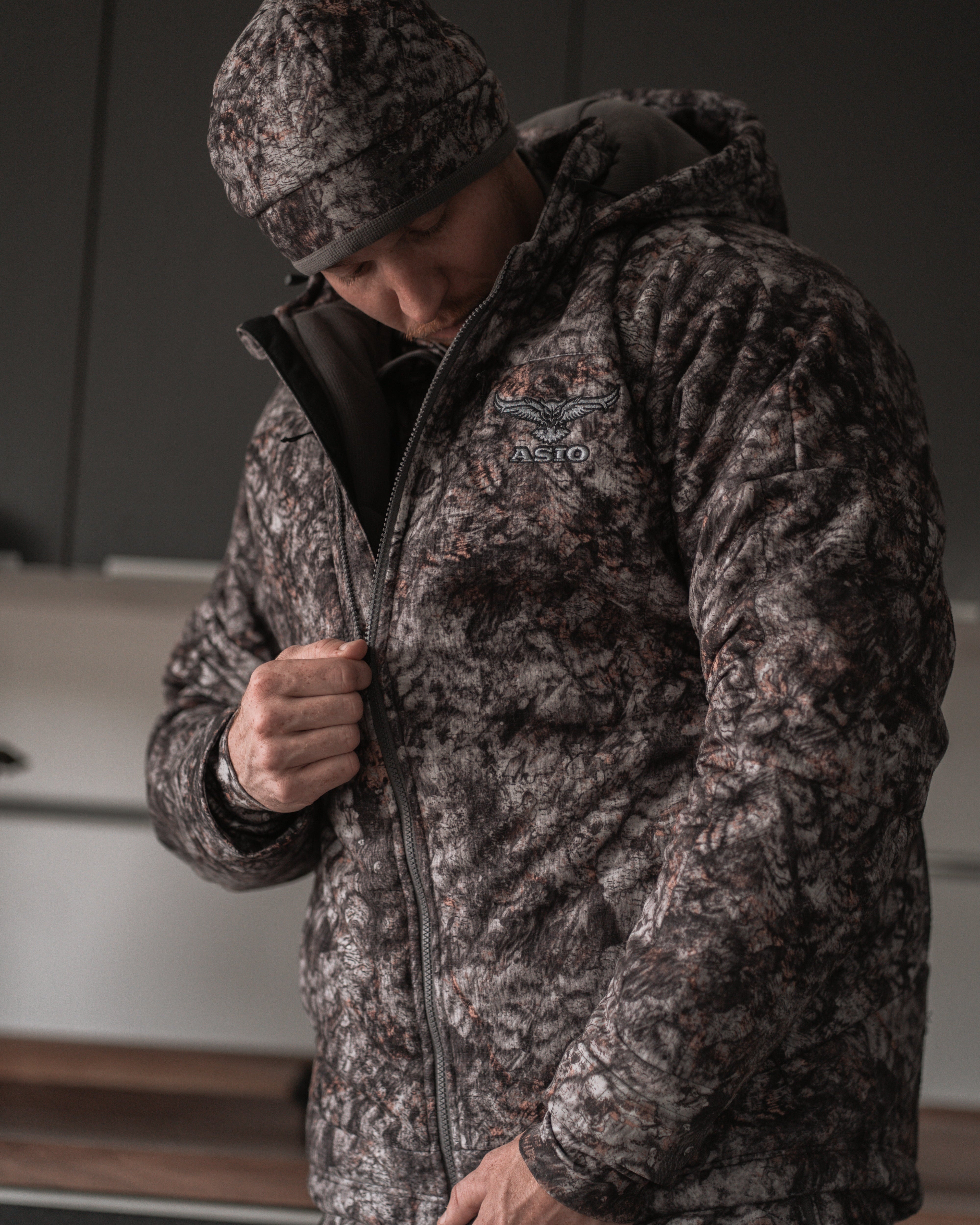 Hunter zipping up ASIO Gear RAPTOR camo heavyweight hooded jacket indoors before cold-weather hunt