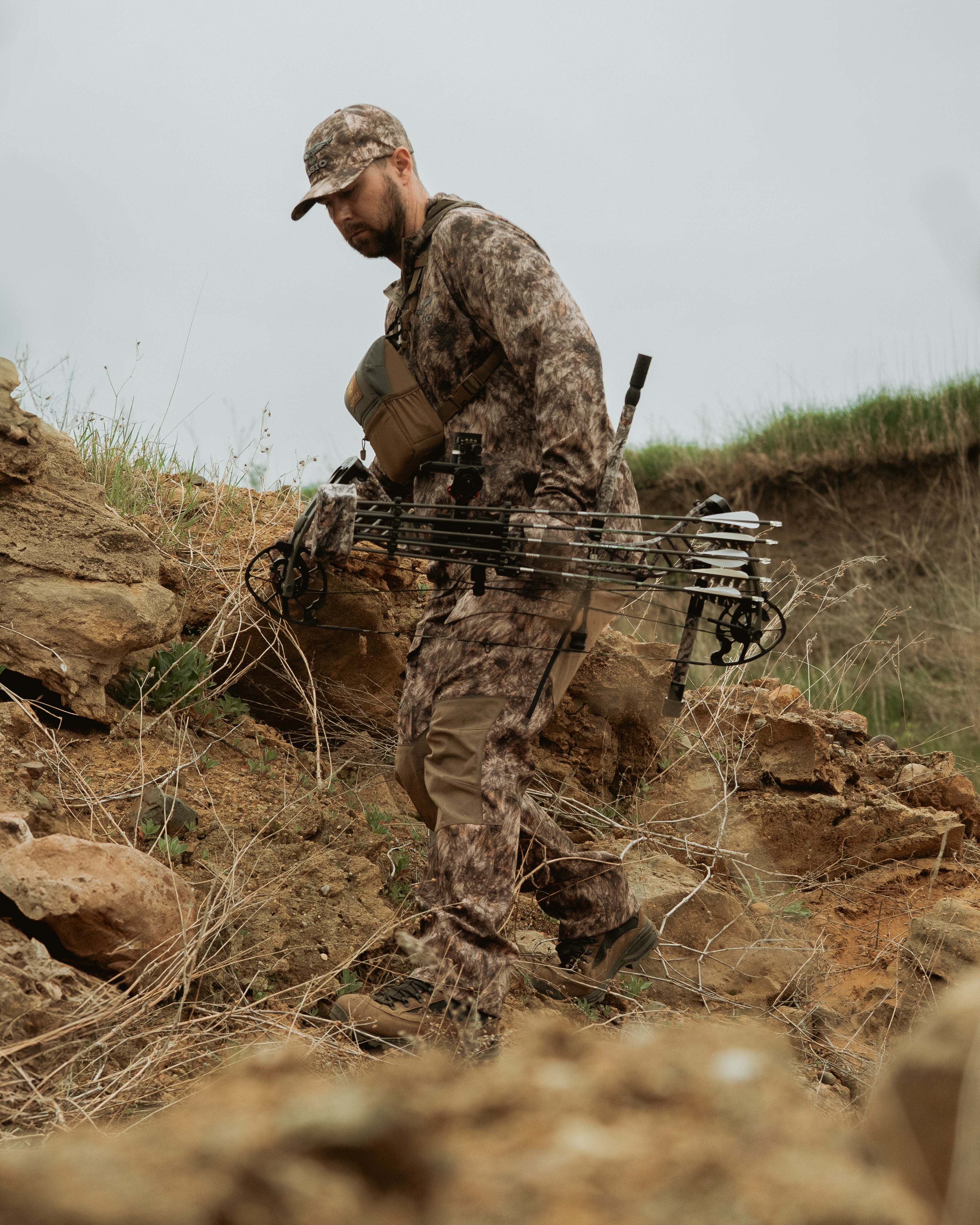 Bowhunter wearing ASIO Gear Midweight Bomb Pant in RAPX camo walking through rugged terrain with mobile hunting setup