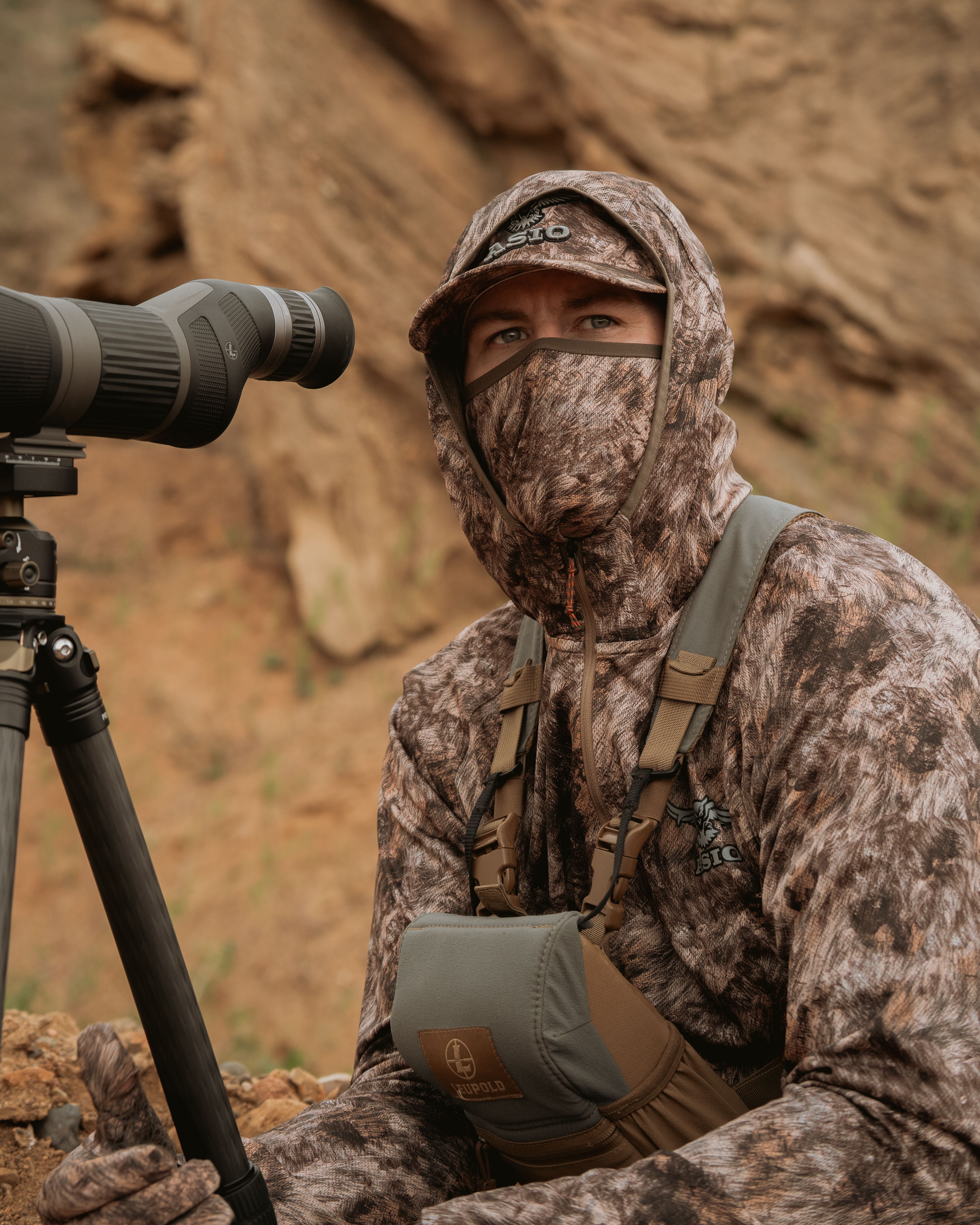 Hunter behind spotting scope in rugged terrain wearing ASIO RAPX Midweight Hoodie and hat.