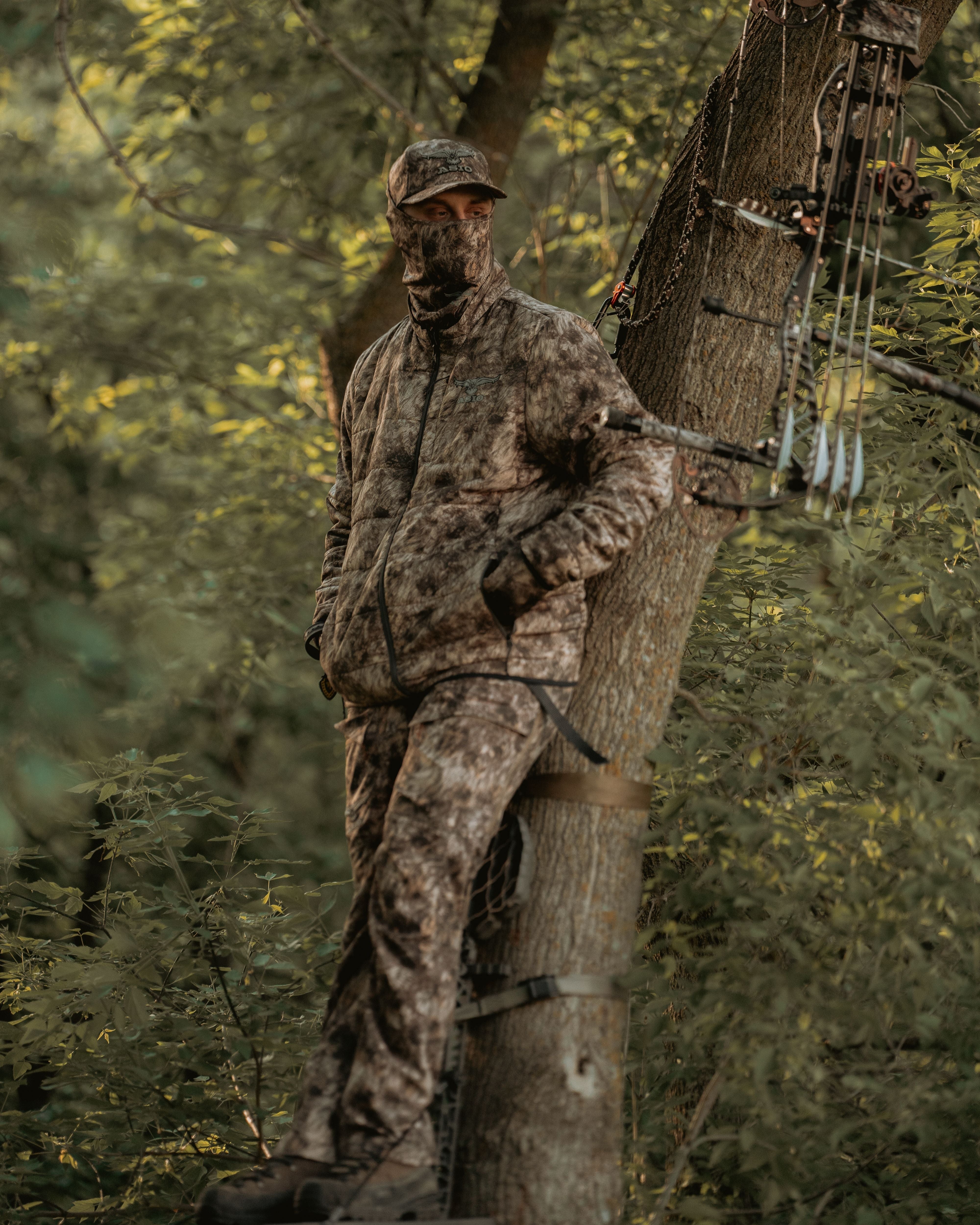 Hunter in ASIO RAPX camo midweight puffy jacket standing in treestand with compound bow and full layering system