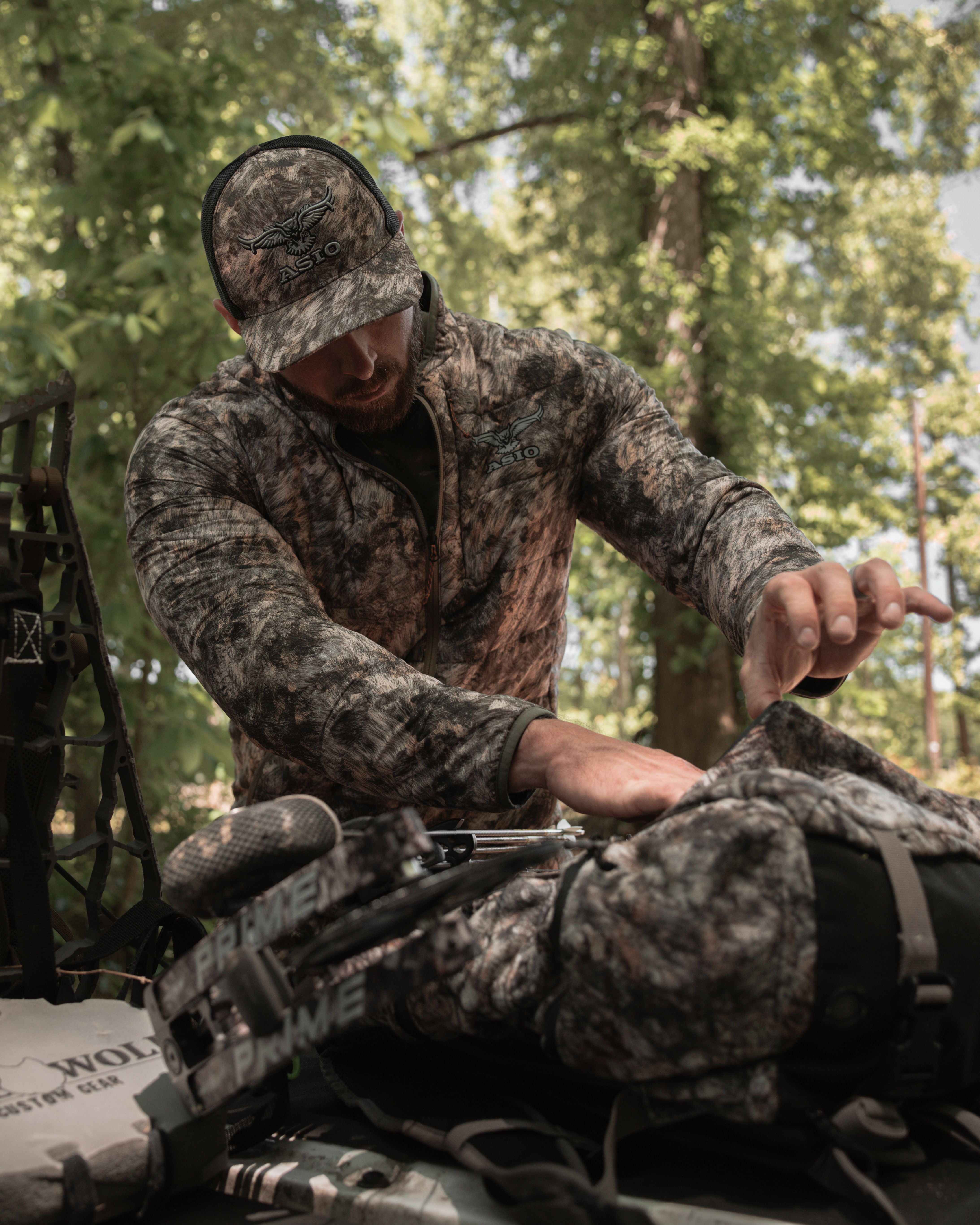Hunter wearing ASIO RAPX camo midweight puffy jacket organizing hunting gear and bow from treestand pack setup
