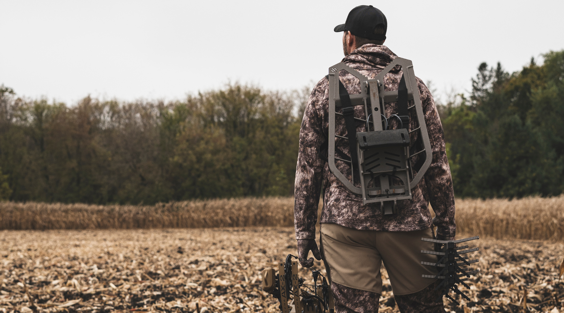 Bowhunter walking through a harvested field wearing ASIO Gear, carrying a bow and pack while scouting whitetail habitat.