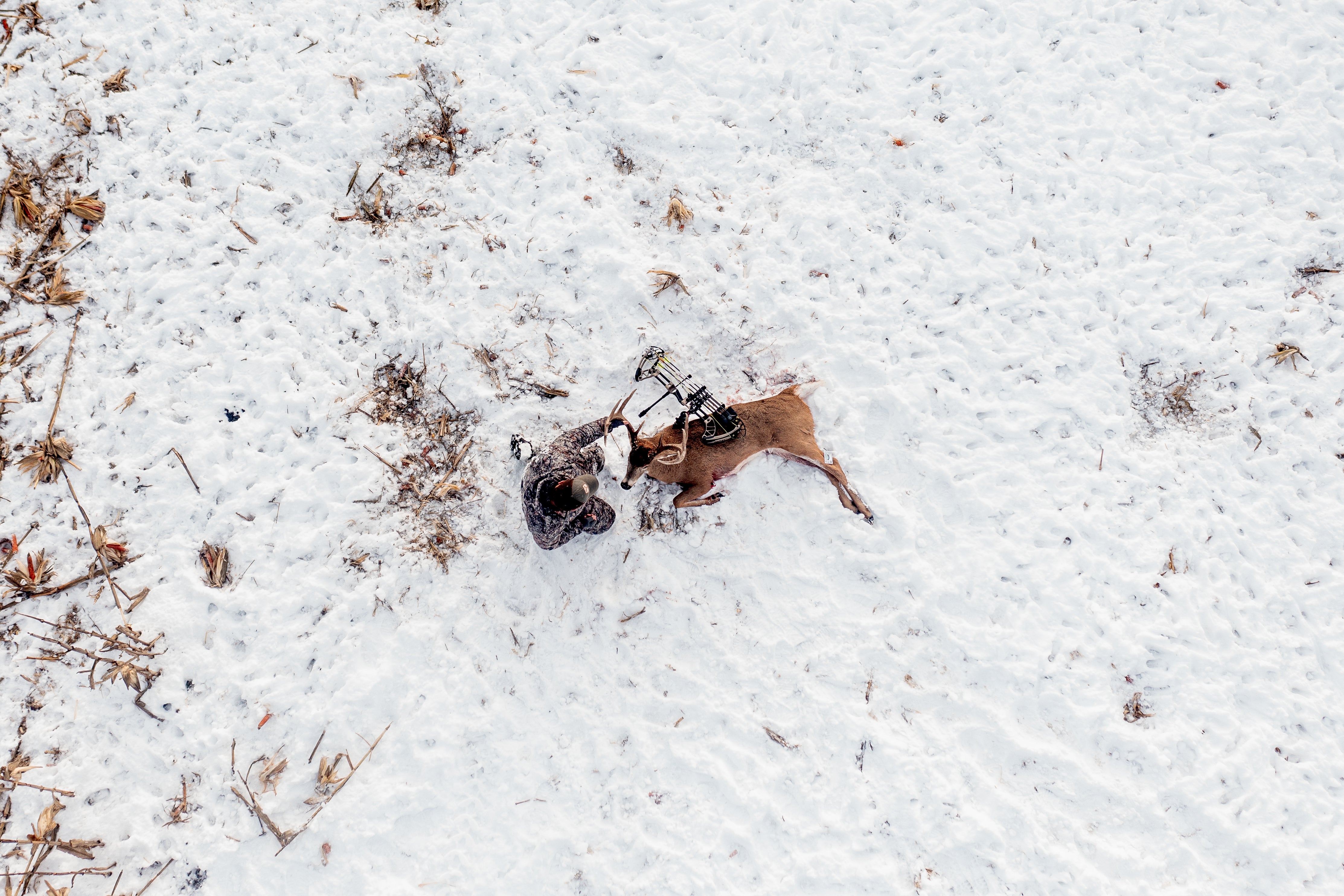 Bowhunter wearing ASIO Gear late-season camo kneeling beside harvested whitetail deer in snowy field