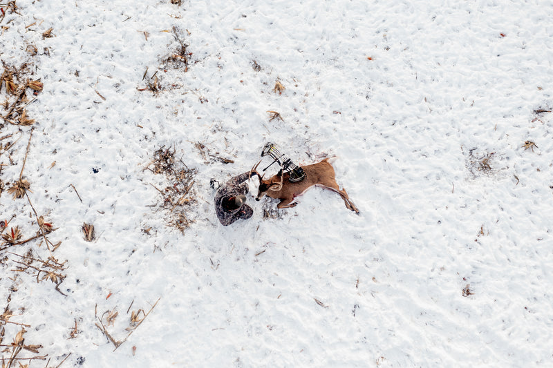Bowhunter wearing ASIO Gear late-season camo kneeling beside harvested whitetail deer in snowy field