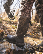A bowhunter wearing ASIO Gear’s Whitetail Pro Boot steps through a shallow creek in late-season woods, shown with ASIO’s Raptor camo pants and a compound bow in hand.